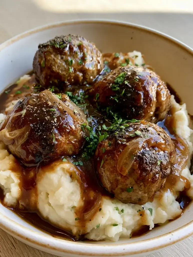 Salisbury Steak Meatballs resting on a bed of fluffy creamy white mashed potatoes with herbs.